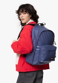 Granatowy plecak z materiału, z przednią kieszenią, regulowanymi paskami i uchwytem do noszenia na górze, noszony na czerwonej kurtce przez osobę.