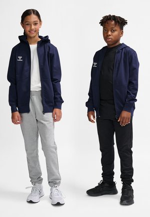 Marineblå hættezipjakke med logo, grå sweatpants og hvide sneakers til venstre; sort skjorte og bukser med sorte sneakers til højre.