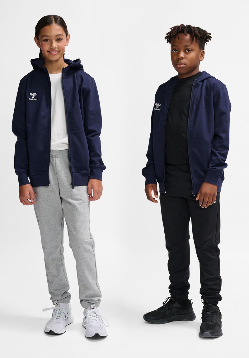 Marineblaues Kapuzensweatshirt mit Reißverschluss und Logo, graue Jogginghose und weiße Sneakers links; schwarzes Shirt und Hose mit schwarzen Sneakers rechts.