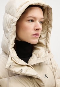 Beige pūkains jakas ar lielu kapuci, ar rāvējslēdzēju priekšpusē un augstu apkakli. Uz jakas krūtīm ir zīmols, un tai ir spīdīga, quiltēta tekstūra.
