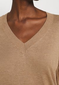 Lichtbruine gebreide trui met een V-hals, voorzien van een zachte textuur en geribbelde accenten langs de halslijn.