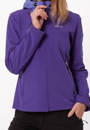 Henkilö, joka pukeutuu violettiin vetoketjulliseen hupulliseen takkiin, säätää vetoketjua kauluksen lähellä ja jolla on mustat housut, toinen käsi taskussa.