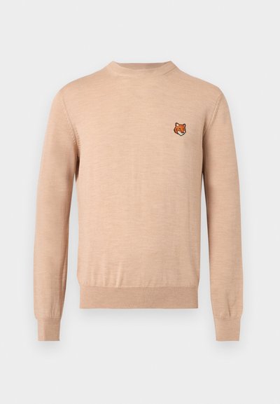 Pull beige à manches longues avec col rond, poignets et ourlet côtelés, arborant un petit emblème de tête de renard orange sur le côté gauche de la poitrine.