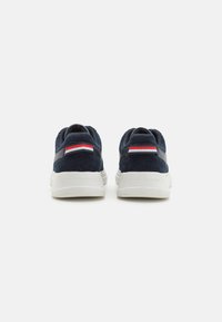 Pantofi sport din suede bleu marin cu talpă din cauciuc albă, având o dublă bandă orizontală în roșu, alb și albastru pe călcâi. Logo-ul este vizibil pe lateral.