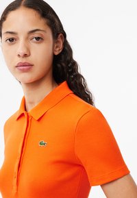 Orange ribbade polo med krage, med en liten grön krokodil-logotyp på vänster sida. Korta ärmar med en figursydd design.