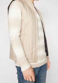 Person som bär en beige vadderad väst över en vit och beige randig tröja tillsammans med blå jeans, som står framför en enfärgad bakgrund.