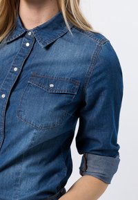 Chemise en denim bleu moyen avec un col à revers, des boutons-pression argentés, deux poches poitrine et des manches retroussées. Texture lisse.