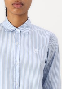 Camicia button-up a righe verticali blu e bianche con colletto curvo e logo ricamato sul lato sinistro. Tessuto dalla texture liscia.