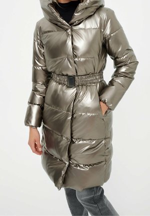 Frau trägt eine silbermetallische, gesteppte Steppjacke mit Kapuze und tailliertem Gürtel, kombiniert mit grauer Hose und schwarzem Rollkragenpullover.