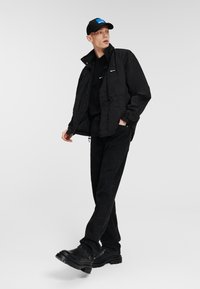 Chaqueta de nylon negra con un cuello alto, combinada con una camiseta negra, jeans negros y zapatos negros brillantes. Completo con una gorra negra con un acento azul.