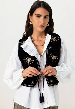 Black velvet vest with gold starburst embroidery, worn over a white ruffled blouse. Decorative ties with tassels accent the design.