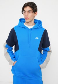 Brezelo in mornarica barvno blokiran pulover s kapuco, s kenguru žepom, vrvico za nastavljanje kapuce in belim Nike logotipom. Narejen iz mehkega materiala, z rebrastimi manšeti.