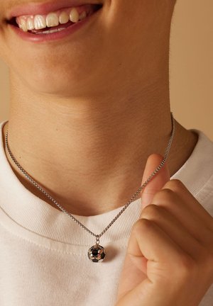 Zilveren ketting met een kleine zwarte en zilveren voetbalsleutelhangertje, geplaatst tegen een lichte stoffen achtergrond.
