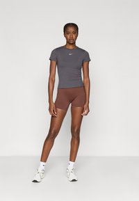 Γκρι εφαρμοστό αθλητικό t-shirt με κοντά μανίκια και λευκό λογότυπο, σε συνδυασμό με καφέ σορτς. Το μοντέλο φοράει λευκά αθλητικά παπούτσια και κάλτσες.