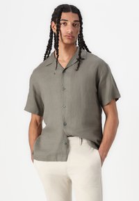 Maglietta di lino verde oliva a maniche corte con colletto a punta e chiusura frontale con bottoni, abbinata a pantaloni beige chiaro.