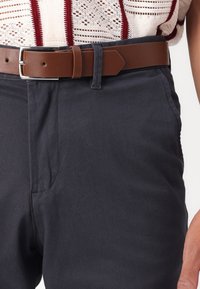 Marineblauwe broek van textuurstof, met zijzakken en een bruine leren riem met een zilveren gesp. Eenvoudig ontwerp, op maat gemaakt.