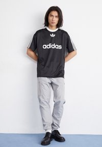 Fekete Adidas póló fehér logóval és gallérral, szürke cargo nadrág cipzáras zsebekkel, és fekete fűzős csizmák egy világos háttéren.