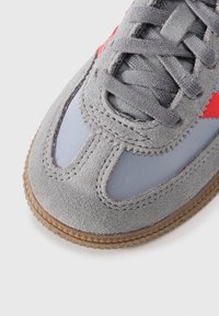 Sneaker en suède gris et synthétique avec des accents rouges, bout rond et semelle en caoutchouc texturé. Offre un design classique à lacets.