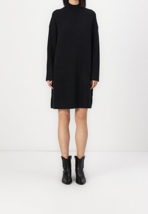 Zwart gebreide jurk met hoge hals, lange mouwen en rechte snit. Draagt zwarte enkellaarzen. Eenvoudig, effen kleur zonder patronen.