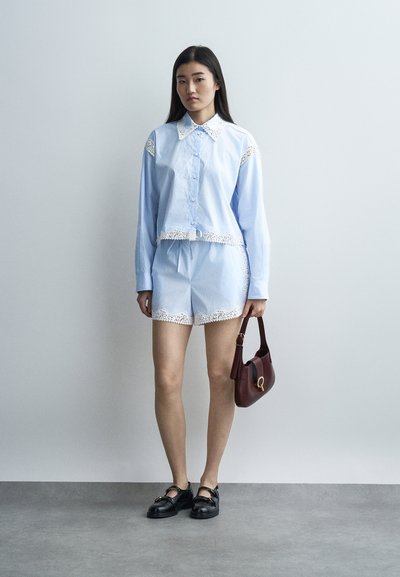 Νεαρή γυναίκα που φορά ανοιχτόγαλάζιο πουκάμισο με κουμπιά και ταιριαστό σορτς με λευκή δαντέλα στο τελείωμα, κρατώντας μπορντό τσάντα, στέκεται σε γκρι πάτωμα.