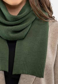 Fular verde tricotat, cu o textură netedă și margini ondulate, așezat în mod casual peste un top negru și o îmbrăcăminte exterioară de culoare deschisă.