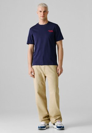 Jeune homme aux cheveux blonds courts portant un t-shirt bleu marine Red Bull Racing, un pantalon beige et des baskets blanches, debout devant un fond uni.