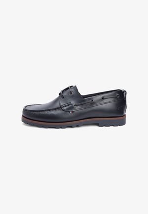 Zapato casual de cuero negro con cuello acolchado, dos ojales para cordones, suela marrón contrastante y detalle de logo en relieve en el lateral.