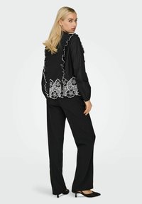 Blouse noire avec détails en dentelle florale blanche, accents à volants et manches longues, associée à un pantalon noir et des escarpins à bout pointu.