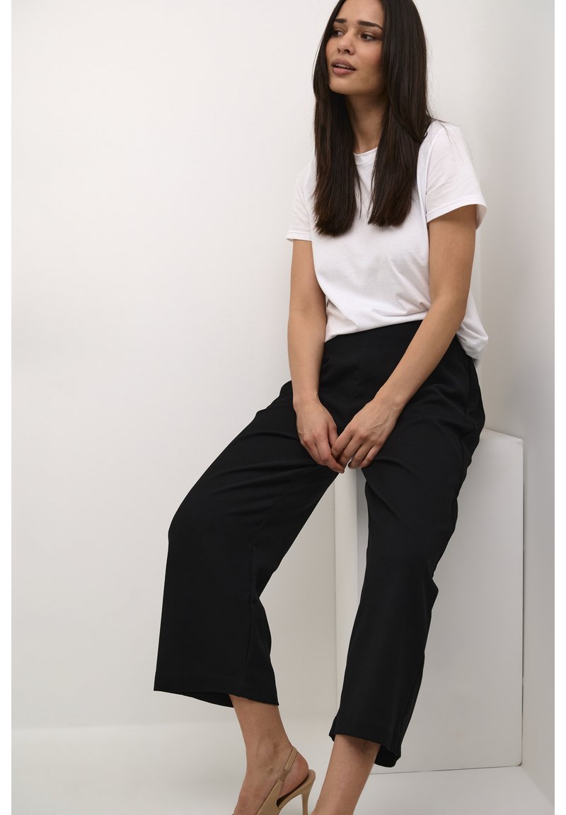 Un t-shirt blanc à manches courtes associé à un pantalon noir à jambes larges. Le modèle est assis pieds nus dans des talons ouverts beiges sur un piédestal blanc.