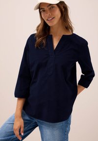 Vrouw in een marineblauwe blouse met lange mouwen, lichtblauwe jeans en een crèmekleurige pet tegen een effen beige achtergrond.