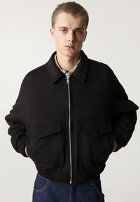 Giubbotto bomber nero con zip frontale, due tasche anteriori, polsini a costine e una texture liscia, abbinato a jeans in denim.