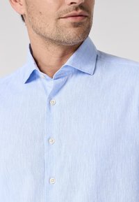 Camicia azzurro chiaro con bottoni, realizzata in tessuto texturizzato, con colletto a punta e bottoni bianchi lungo la pattina frontale.