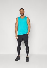 Turkoosi hihaton paita, mustat shortsit, mustat legginsit ja mustat lenkkarit, joissa on valkoinen Nike-logo. Malli seisoo kädet taskuissa.