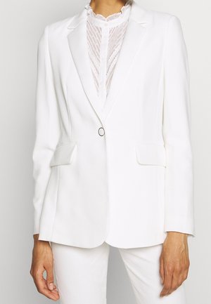 White tailored blazer with single button closure worn over a semi-sheer white lace blouse and white pants.