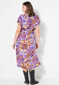 Lila und braun gemustertes Kleid mit Paisleymuster, kurzen Ärmeln, tailliertem Schnitt und knöchellangem Saum, kombiniert mit schwarzen Cowboystiefeln.