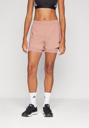 Kvinde iført lyserøde lagdelte atletikshorts, sort sports-BH, hvide sokker med logo og sorte atletiksko, stående mod en ensfarvet baggrund.