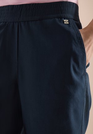 Osoba nosząca ciemnogranatowe spodnie z elastycznym pasem, z ręką w prawym kieszeni i bladoróżową koszulę schowaną do środka.