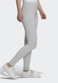 Person i ljusgrå leggings och vita sneakers, stående med ett ben något böjt mot en enfärgad ljus bakgrund.