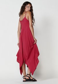 Rotes, fließendes Maxikleid mit dünnen Trägern und asymmetrischem Saum. Hergestellt aus leichtem Material, mit einem Knopfdetail am Rücken. Schwarze Sandalen mit Riemchen.