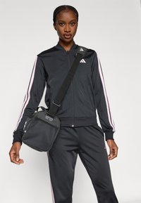 Μαύρη αθλητική φόρμα με ροζ ρίγες, που περιλαμβάνει ζακέτα με φερμουάρ και ταιριαστό παντελόνι. Κρατάει μια μικρή μαύρη τσάντα ώμου από δίχτυ.