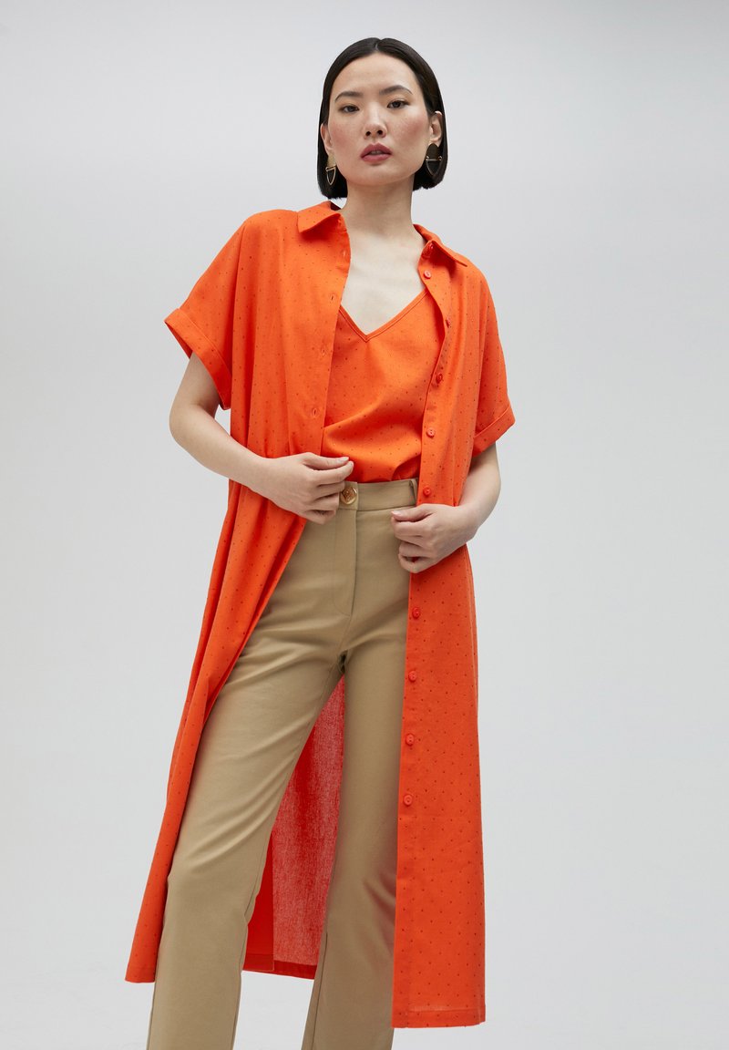 Oranges, knöpfbares, langes Hemd mit kurzen Ärmeln; strukturiertem Stoff. Darunter ein passendes orangefarbenes Tanktop. Beigefarbene, eng anliegende Hose mit einem goldenen Knopf.