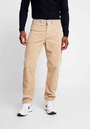 Homme portant un pantalon en velours beige, une chemise noire à manches longues et des baskets blanches avec un logo Nike noir, debout sur un sol blanc uni.