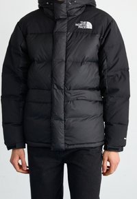 Chaqueta acolchada negra aislante con cierre delantero, capucha y logo. Confeccionada con un material suave y ligero, presenta segmentos de corte cuadrado.