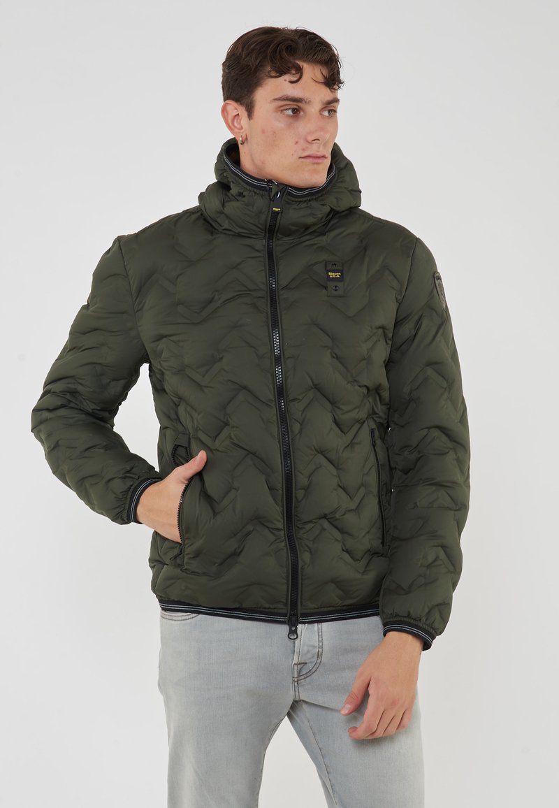 Giacca a vento verde, trapuntata, con colletto alto, zip frontale e polsini elasticizzati. Presenta una patch con logo e tasche laterali. Texture liscia e imbottita.