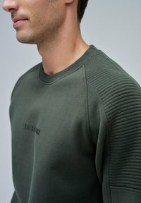 Uomo che indossa un felpa verde oliva con maniche a coste e un sottile logo "SALSA/MVS" sul petto, mostrato dalla gola fino a metà torace.