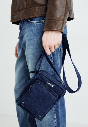 Personne portant un jean bleu et une veste en cuir marron tenant un petit sac à bandoulière Levi's bleu foncé par sa sangle.