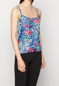 Femme portant un débardeur bleu avec des motifs floraux et feuillages colorés, associé à un pantalon noir, visible de la poitrine jusqu'aux hanches sur un fond blanc.