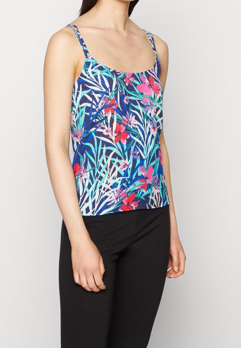 Femme portant un débardeur bleu avec des motifs floraux et feuillages colorés, associé à un pantalon noir, visible de la poitrine jusqu'aux hanches sur un fond blanc.