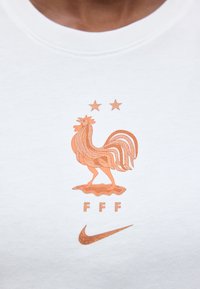 Maglia sportiva bianca con emblema di gallo in bronzo, due stelle sopra, lettere "FFF" sotto e logo Nike in bronzo sul fondo.