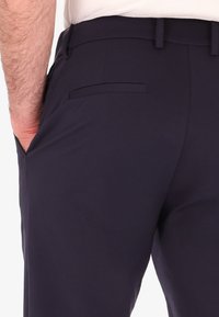 Pantaloni blu navy con una trama liscia, presentano una sola tasca posteriore e passanti per la cintura, la vestibilità su misura è visibile dalla vista laterale.